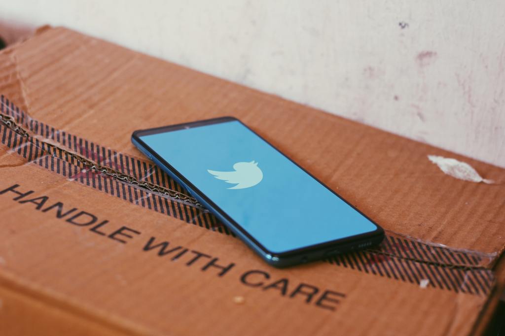 A phone showing a Twitter logo on top of a cardboard box marked 'Handle With Care'.