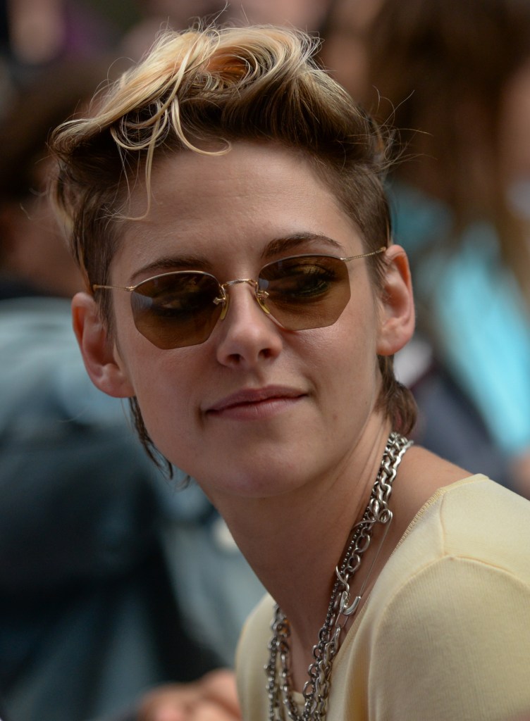Kristen Stewart, smiling slightly, with short messy blonde hair, cool sunglasses and thick silver chains around her neck.