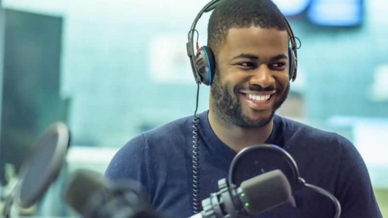 Ben Hunte wears headphones in a radio studio and smiles to someone off camera.