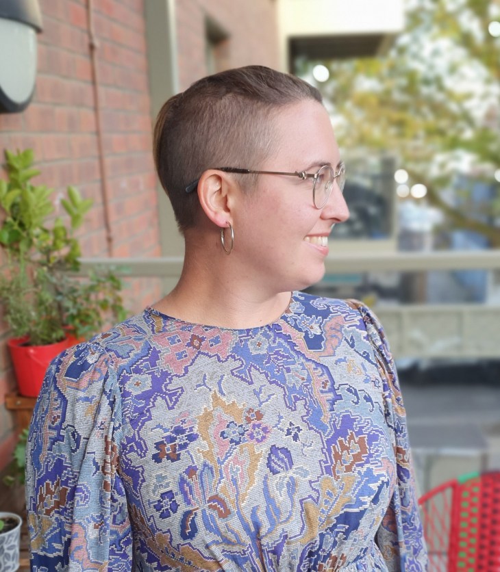 A woman with a shaved topknot hairstyle, outdoors on a balcony with plants in the background, smiling and looking to the right, wearing a blue floral patterned dress and gold hoop earrings.