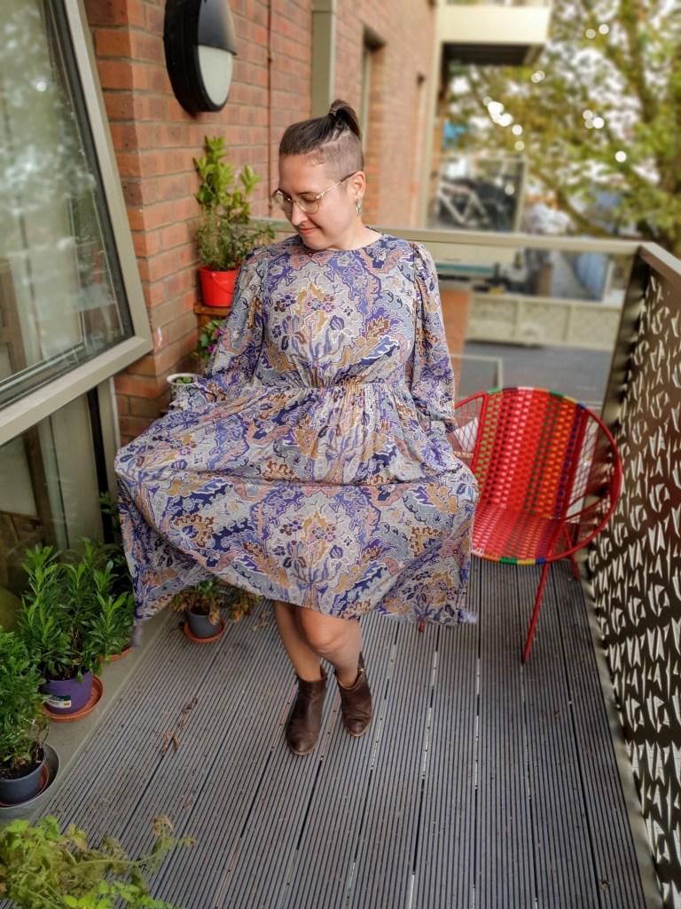 A young woman on a balcony with some pot plants, looking downwards and smiling as she holds out the skirt of her blue patterned dress to show that it has pockets.