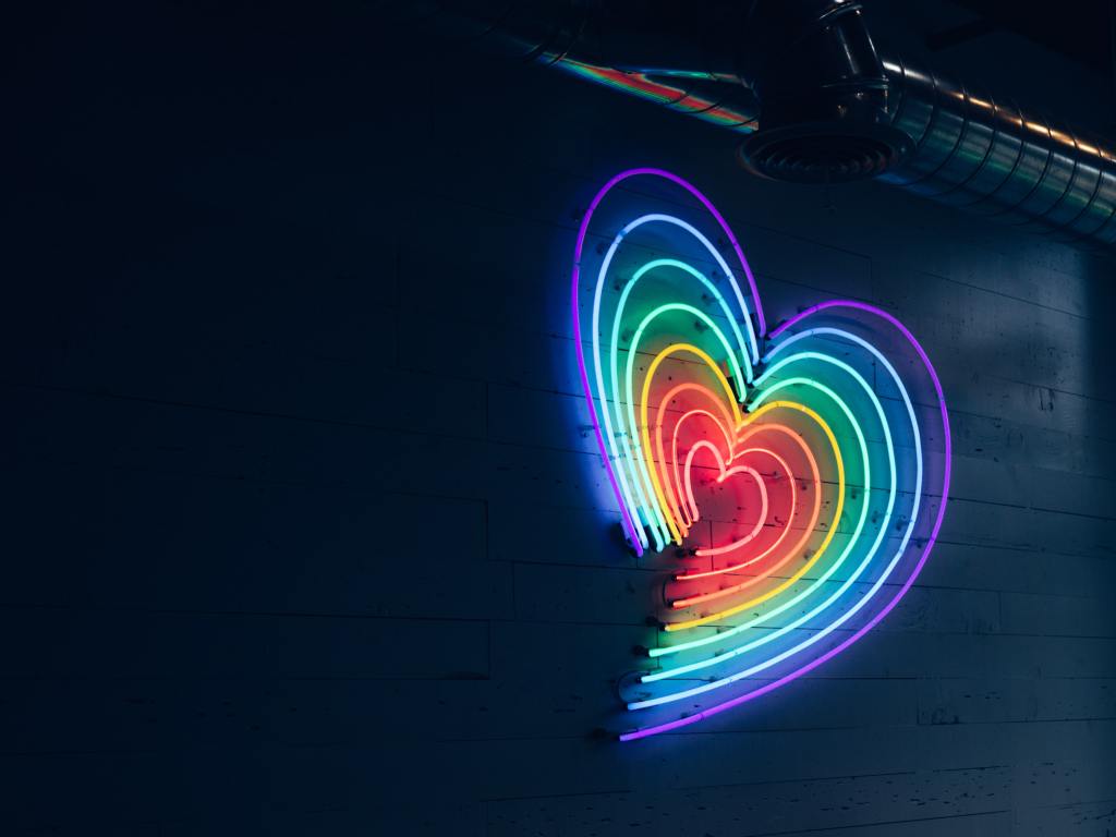 A neon rainbow coloured heart sign glows against a dark wall.