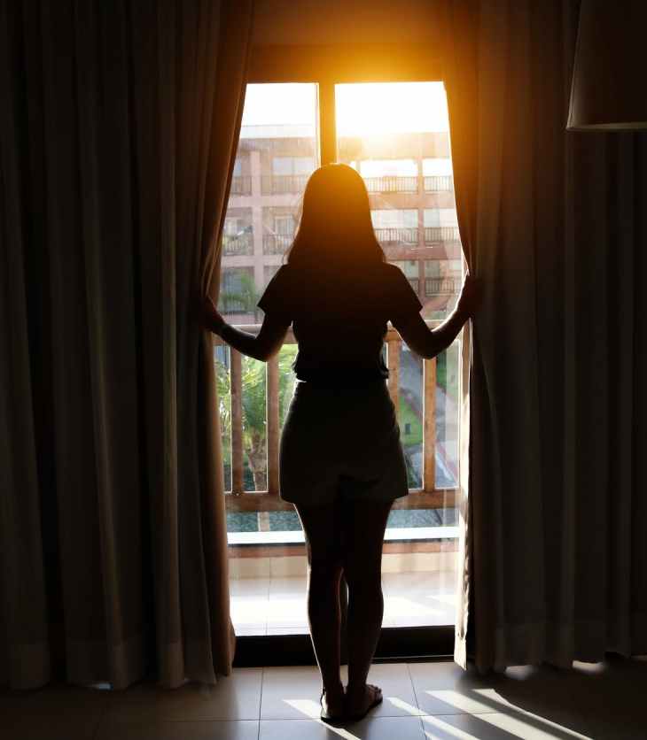 A woman looks through some open curtains to sunshine. She has her back to us and is silhouetted against the light.