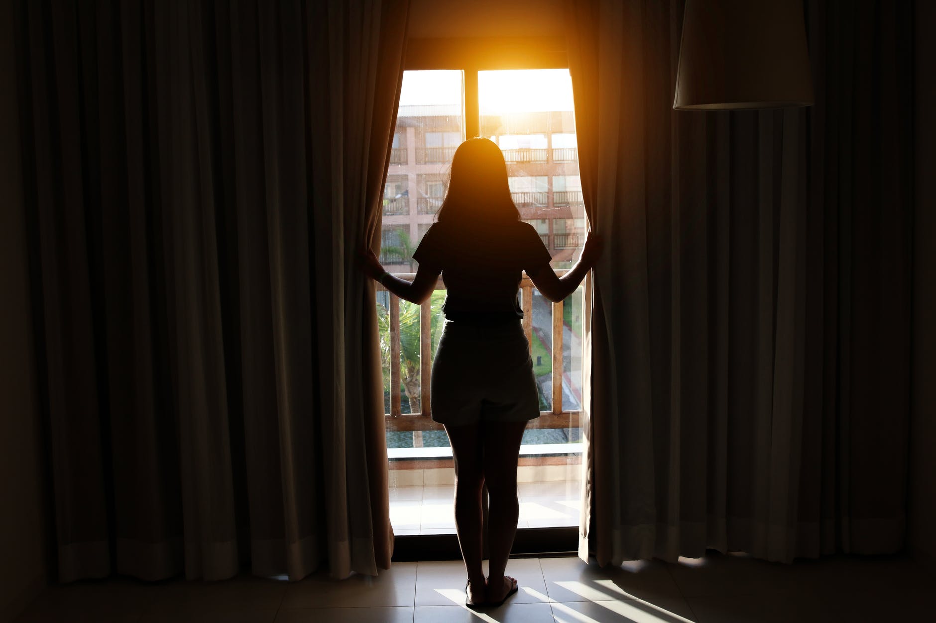 A woman looks through some open curtains to sunshine. She has her back to us and is silhouetted against the light.