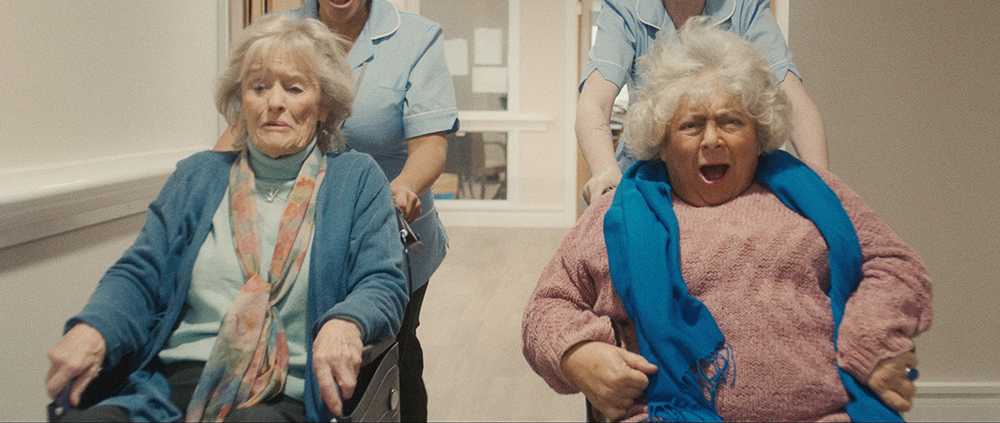 Virginia McKenna and Miriam Margolyes sit in wheelchairs side by side, smiling as they're pushed at high speed down a corridor by nurses in blue uniforms