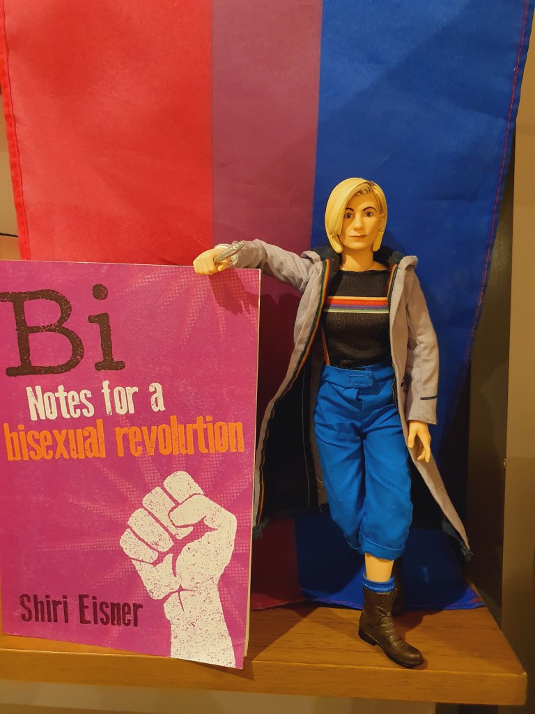 An action figure of Jodie Whittaker as Doctor Who stands on a wooden shelf, leaning on a copy of Bi - Notes on a Bisexual Revolution by Shiri Eisner, with a bisexual pride flag as a background.