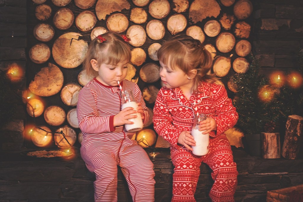 Two little white girls in red and white onesie pyjamas sit against a background of sawn-off logs and Christmas lights, drinking milk from bottles using red and white straws.