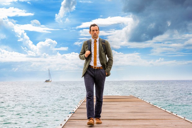 Neville Parker (Ralf Little) looks perplexed as he walks along a jetty towards the camera, with a big blue sky behind him.